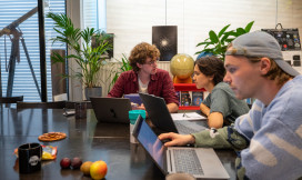 Drie studenten zitten aan een tafel, twee zijn in gesprek en de ander werkt op zijn laptop.