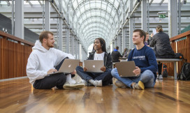 Drie studenten zitten op de grond en zijn in gesprek. Ze werken op hun laptops.