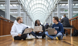 Drie studenten zitten op de grond en zijn in gesprek. Ze werken op hun laptops.