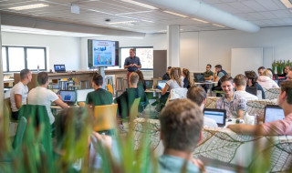 Klaslokaal met studenten achter laptops en een docent voor een groot beeldscherm