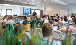 Klaslokaal met studenten achter laptops en een docent voor een groot beeldscherm