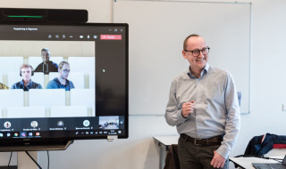 Een beeldscherm met daarop studenten die online deelnemen aan een les en een docent naast het beeldscherm