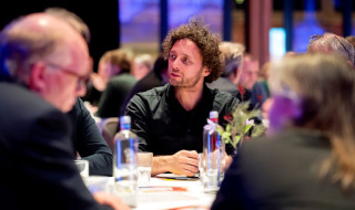 Man met krullend haar en zwart hemd aan een ronde tafel in gesprek met andere volwassenen