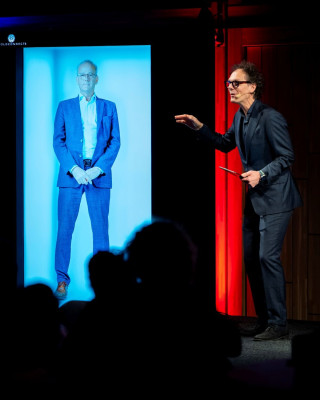 Een man als holografische illusie in een box en een man op het podium die beiden publiek toespreken