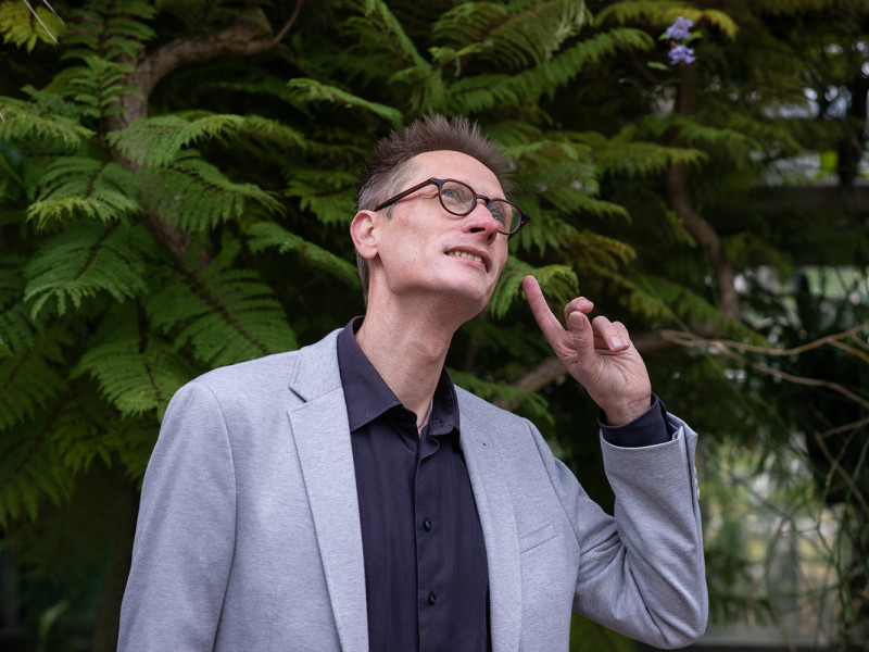 Bob Planqué wijst naar zijn oor om aan te geven dat hij luistert naar vogelgeluiden, op de achtergrond een boom