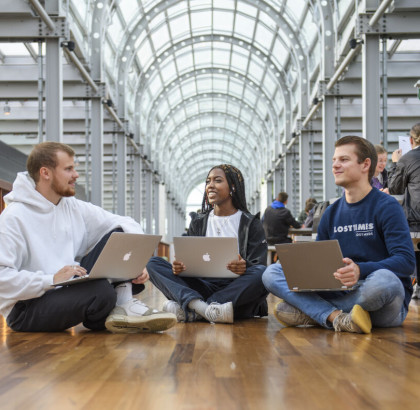 Drie studenten zitten op de grond en zijn in gesprek. Ze werken op hun laptops.