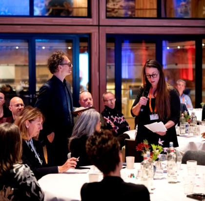 Een vrouw staat met microfoon en leest van papier voor aan publiek dat aan ronde tafels zit.